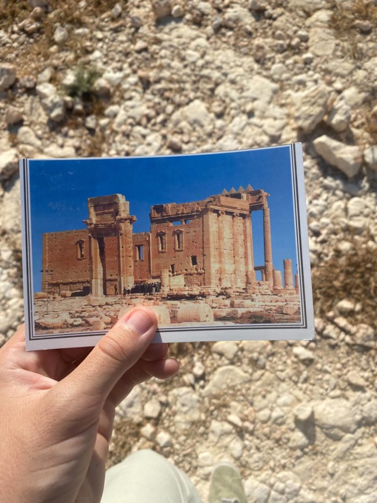 A person visiting Palmyra, holding up a picture of a destroyed building in the desert.