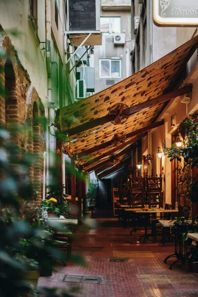 Narrow covered alley in Exarchia with café tables, wooden chairs, warm lights, and close brick and plaster walls on both sides.
