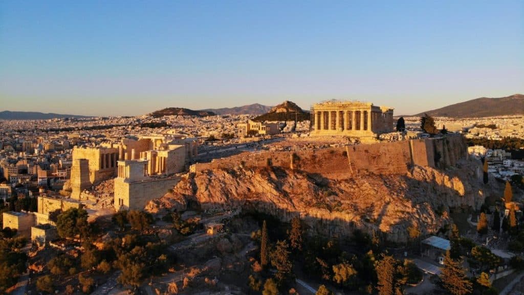 A panoramic view of the Acropolis in Athens during sunset, highlighting the iconic Parthenon and surrounding ancient ruins with a vibrant cityscape background.