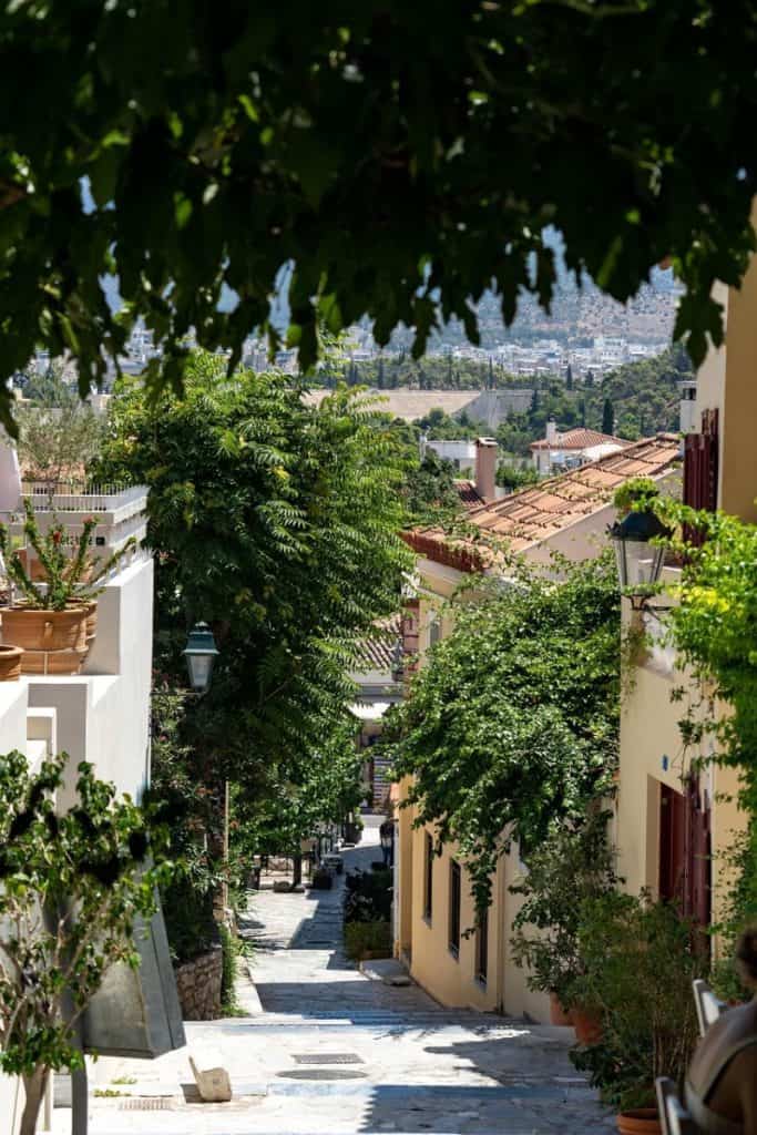 Narrow pedestrian street in Anafiotika lined with pastel houses, potted plants, leafy trees, and a distant view of Athens in the background.
