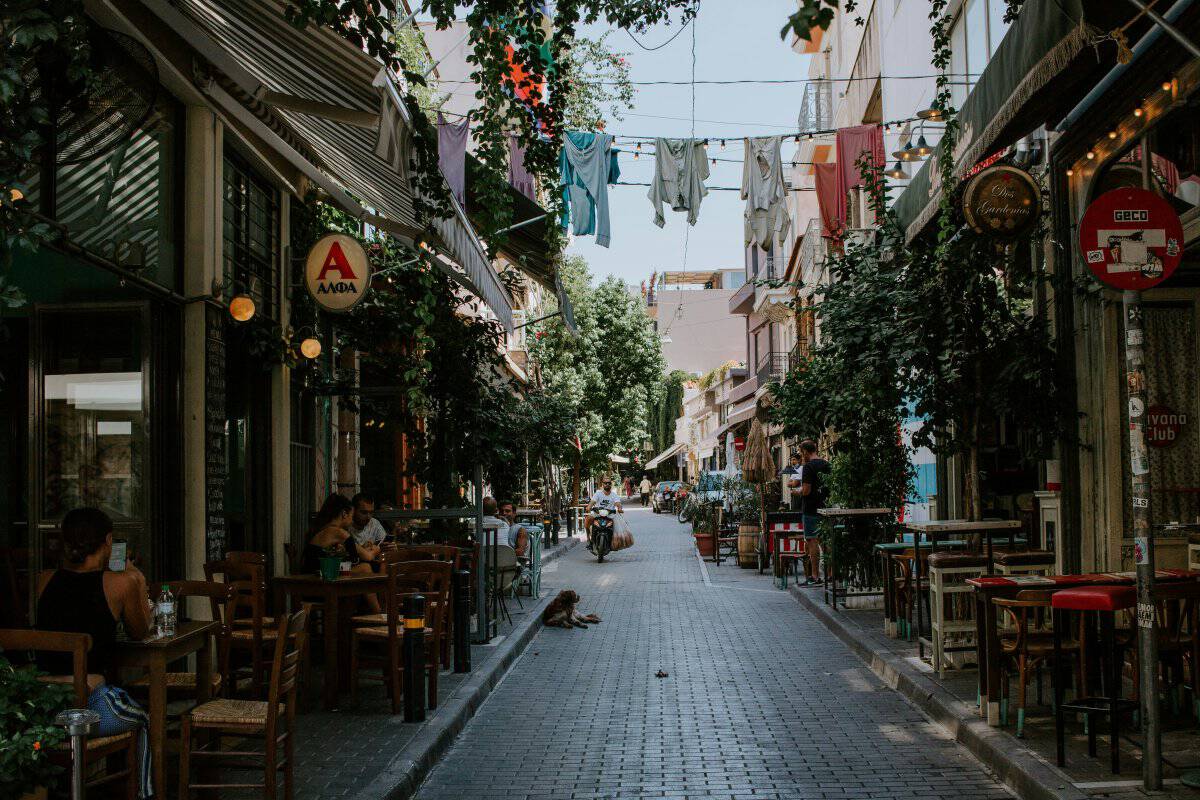 A narrow pedestrian alley in central Athens lined with shops and street signs. Metal awnings jut out from the shopfronts on both sides, while overhead wires and hanging flags cross above the lane. The scene feels distinctly urban, artistic, and slightly hidden, capturing the gritty charm of an old Athens side street.