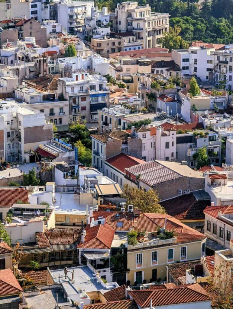 Aerial view of a dense Athens neighbourhood with closely packed apartment buildings, red-tiled roofs, rooftop terraces, balconies, and small patches of greenery between the buildings.
