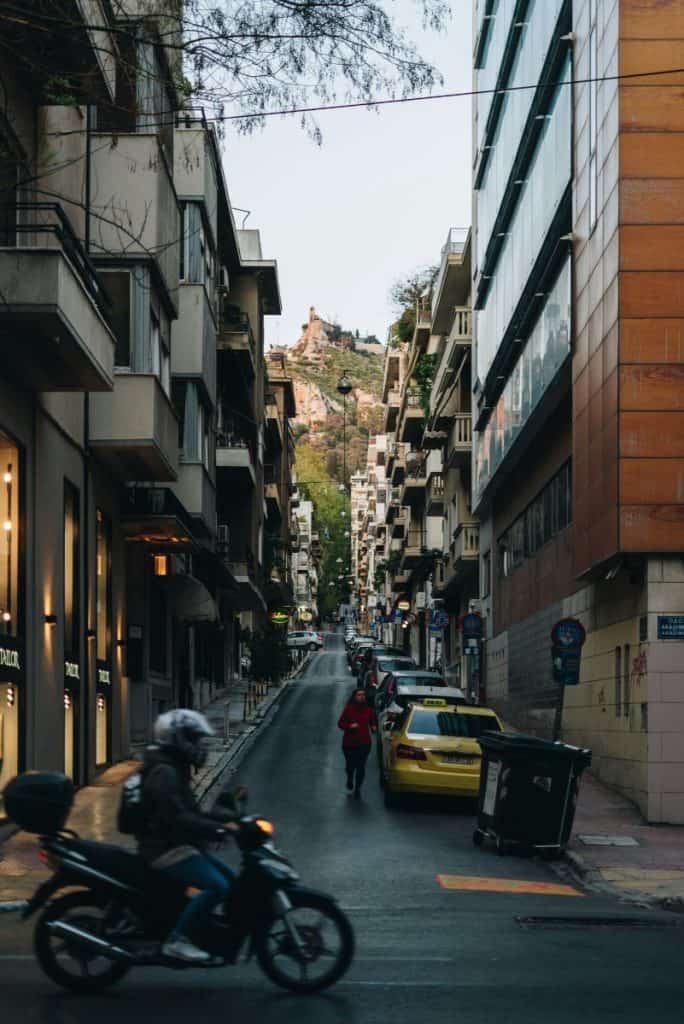 Steep city street in Kolonaki lined with tall apartment buildings, parked cars, a passing motorbike, and Lycabettus Hill rising in the distance.