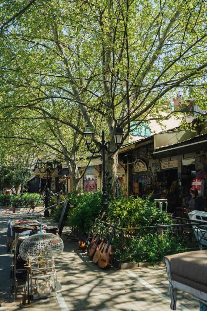 Shaded Monastiraki street with tall trees, outdoor café seating, vintage furniture, hanging guitars, birdcages, and leafy plants along the pavement.
