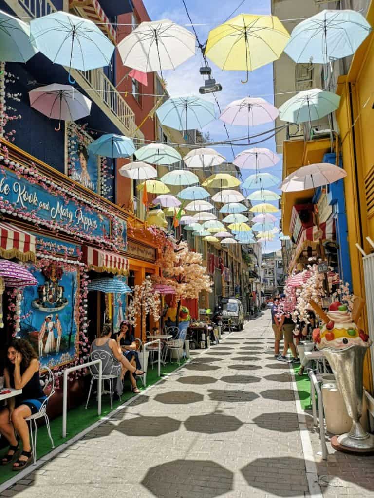 Colourful pedestrian alley in Psirri decorated with hanging pastel umbrellas, bright café facades, outdoor tables, and people sitting and walking.