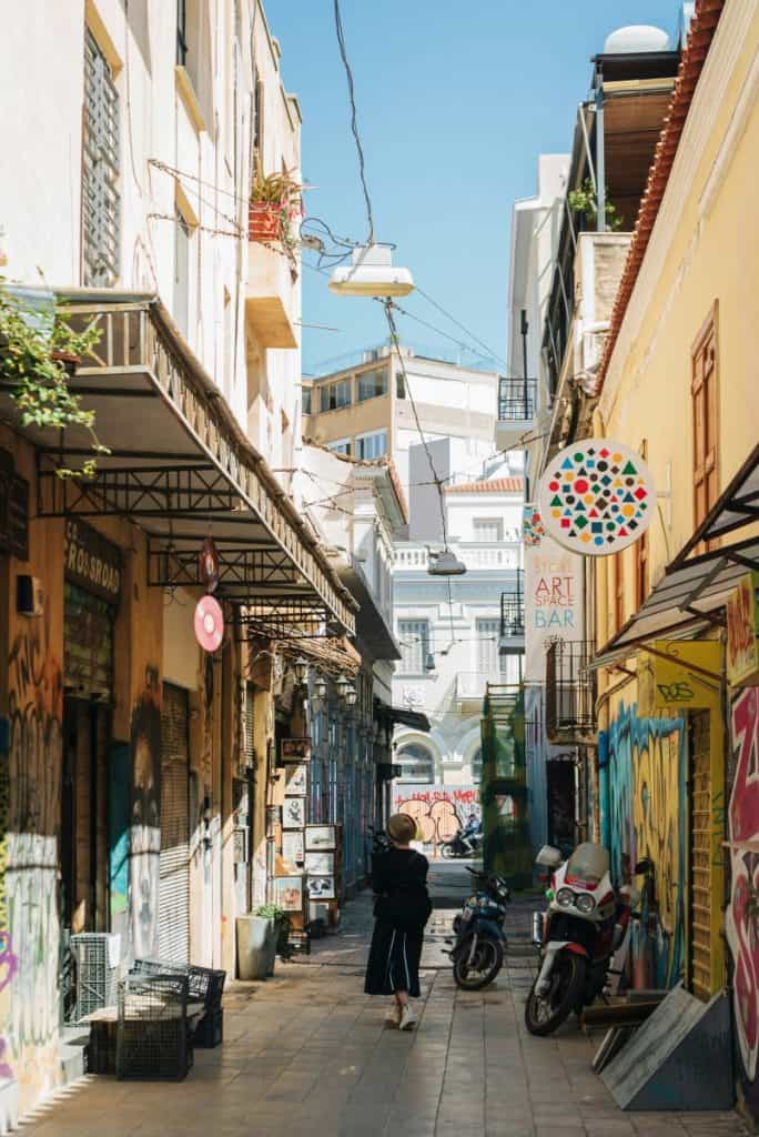 Narrow graffiti-lined alley in Psirri with parked scooters, shuttered storefronts, hanging signs, balconies, and a person walking down the street.
