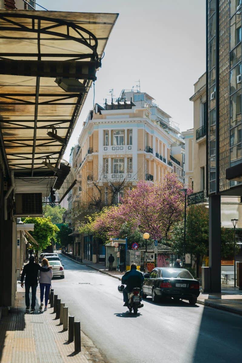 Sunny street near Syntagma with a pale neoclassical building, flowering trees, pedestrians, parked cars, and a motorbike driving through the centre.
