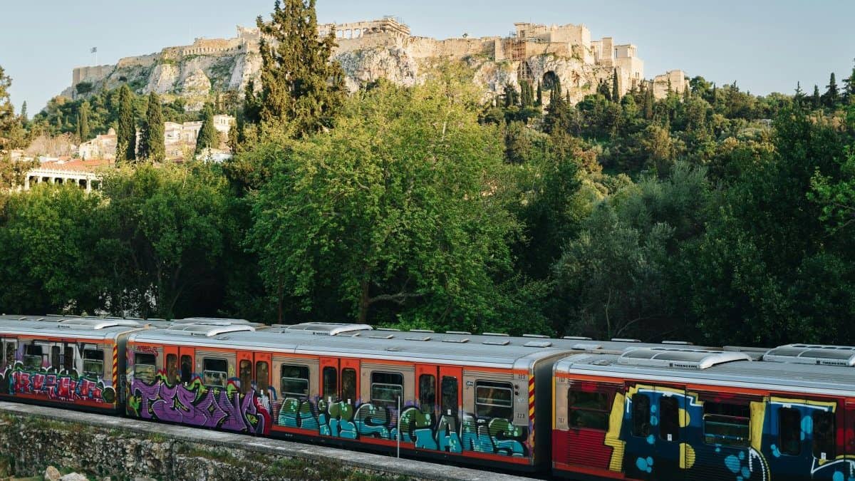 Graffiti-covered train passing through central Athens below the Acropolis, with green trees in the foreground and the ancient hilltop ruins above.
