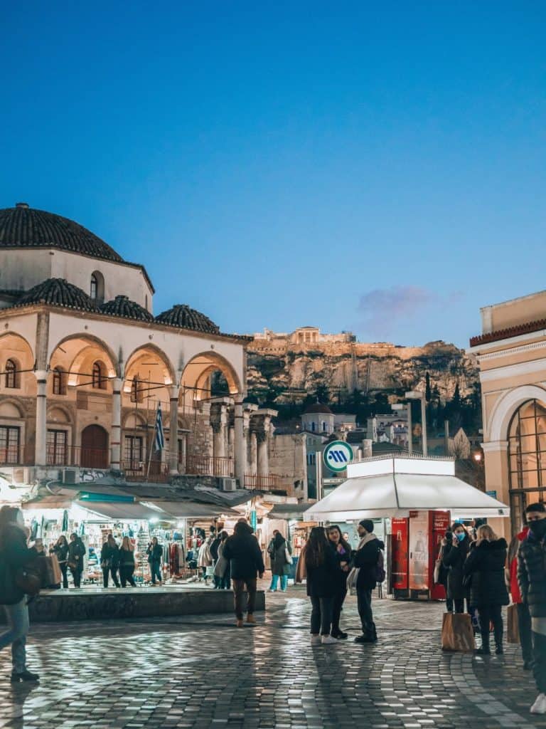 Evening scene in Monastiraki Square with pedestrians, market stalls, the domed Tzistarakis Mosque, and the Acropolis lit up on the hill behind.
