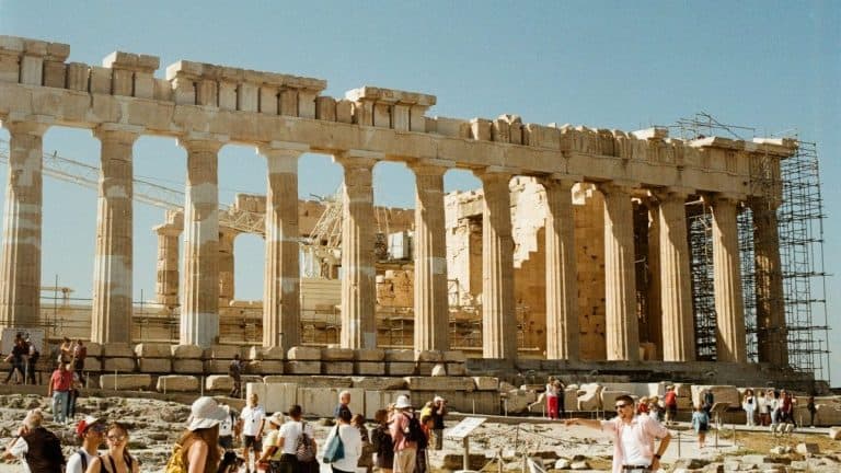 The image features tourists visiting the iconic Parthenon, an ancient Greek temple on the Acropolis.
