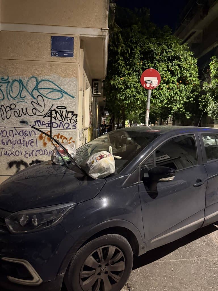 Nighttime street scene in Athens showing a dark compact SUV parked partly on the sidewalk beside a graffiti-covered building. Two plastic trash bags have been left on the car’s windshield with the wipers lifted upright, while a red no-entry traffic sign and orange trees line the narrow residential street in the background.