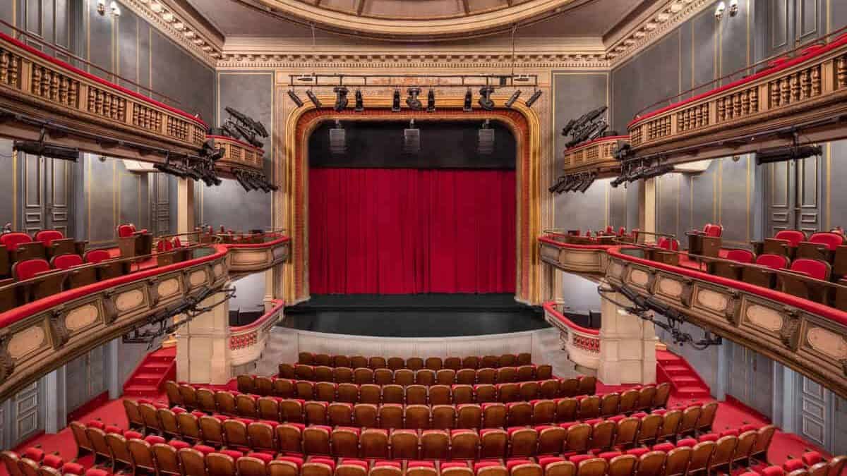 The National theatre interior, seen from the back of the auditorium facing the stage. The space is very symmetrical, with rows of red and brown seats on the ground floor and curved balconies on both sides, all lined with red velvet. The stage is framed by an ornate arch and closed with a deep red curtain. Above, there is a large domed ceiling with decorative painted panels and classical detailing, which gives the whole room a very grand, historic feel.
