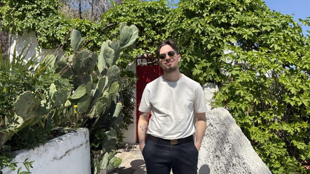 A photo of Nick, owner of lifessmallworldsbig.com in front of a red door covered with green plants in the Anafiotika neighbourhood in Athens.