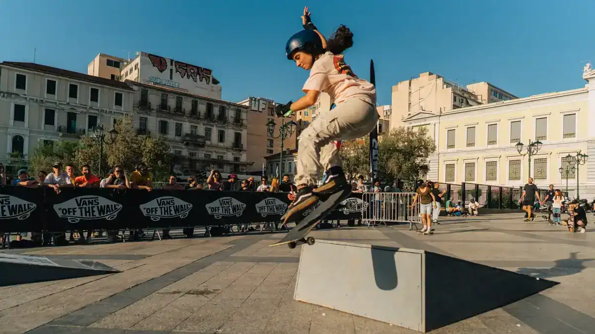 Young skateboarder performing a trick over a metal ramp during a Vans skateboarding event in central Athens, with spectators watching from behind barriers in a sunny urban square.