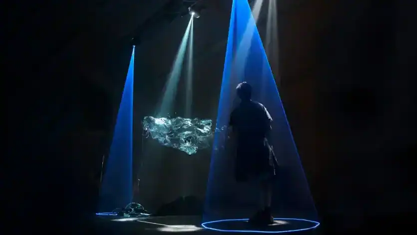 Silhouetted visitor standing beneath blue spotlights inside a dark immersive art installation in Athens, with glowing circular floor lights and floating abstract digital visuals suspended in the air.