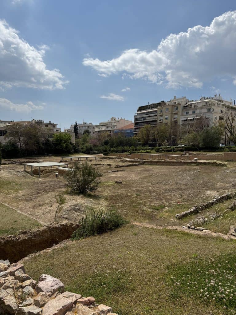 Wide view of the archaeological remains at Aristotle’s Lyceum in Athens, with low stone foundations and dry grassy ground stretching across an open excavation area. Modern apartment buildings and trees surround the site under a bright blue sky with large white clouds.