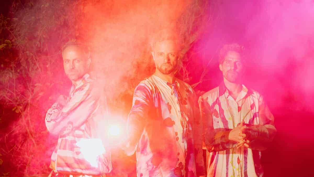Three performers wearing colorful patterned outfits surrounded by thick pink and orange smoke during a vibrant outdoor performance at the Athens Jazz Festival at night.