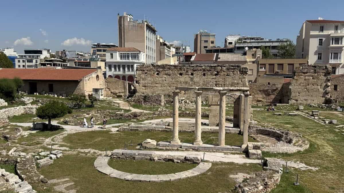 Elevated view inside Hadrian’s Library in central Athens, showing ancient stone ruins and a small rectangular structure with four tall marble columns standing in the middle of a grassy archaeological site. Old stone walls and modern city buildings rise in the background beneath a clear sky.