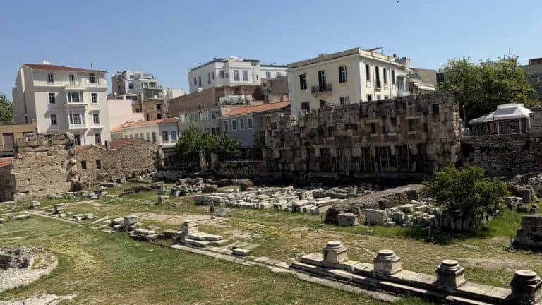Interior view of Hadrian’s Library with scattered ruins, broken columns, and sections of ancient stone walls spread across a large open archaeological area. Multi-story residential buildings surround the site, contrasting the ancient remains with the modern city.