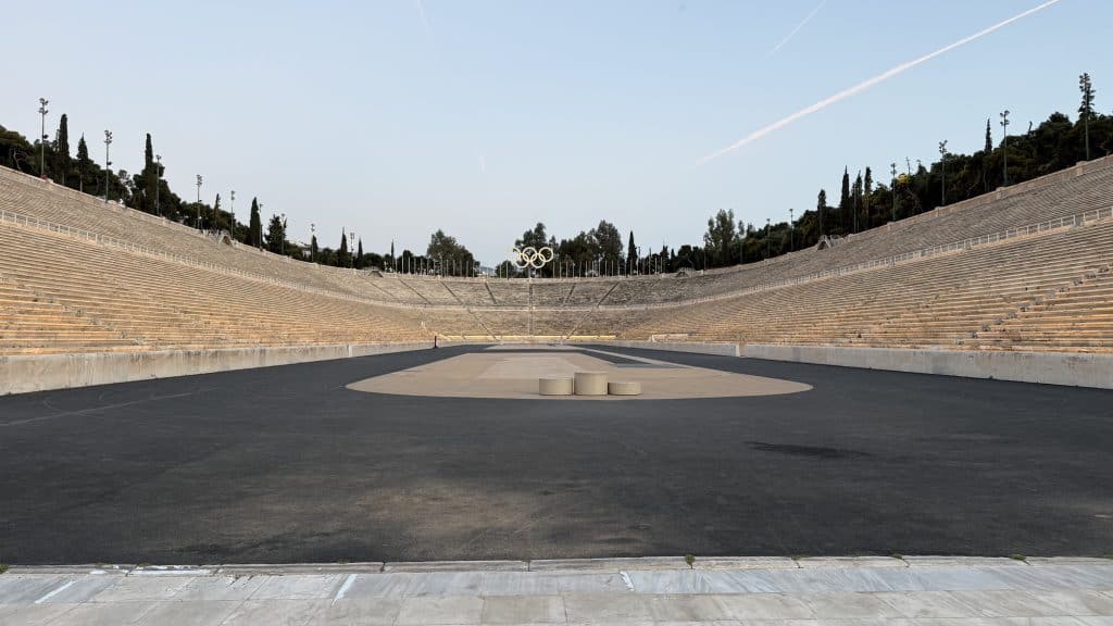 Symmetrical view from the center of the Panathenaic Stadium in Athens, looking across the dark running track toward long rows of pale marble seating curving around both sides. The Olympic rings stand in the distance at the far end of the stadium.