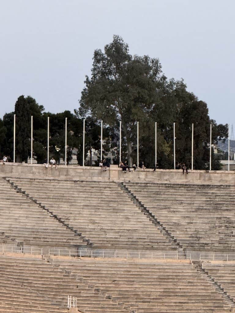 Close-up view of the upper marble seating area of the Panathenaic Stadium, with several people sitting and standing along the top edge near a row of tall flagpoles and large trees in the background.
