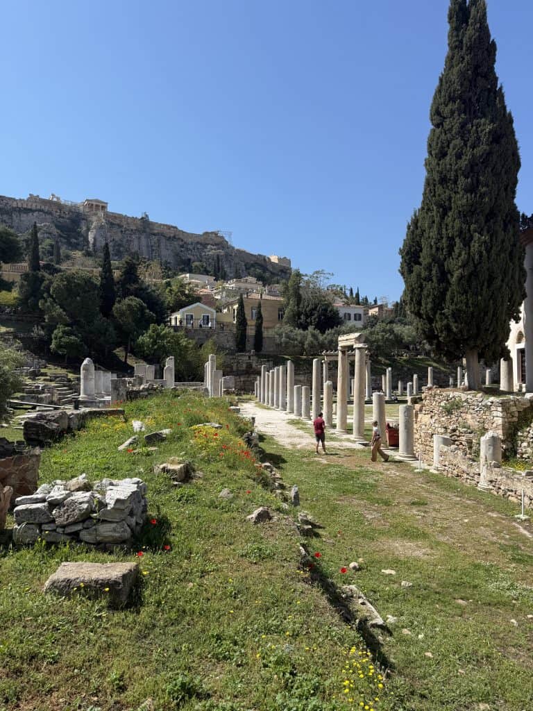 View inside the Roman Agora of Athens with a pathway lined by ancient marble columns leading toward the Acropolis hill in the distance. Wildflowers, grass, stone ruins, and a few visitors walking through the site fill the foreground under bright sunlight.