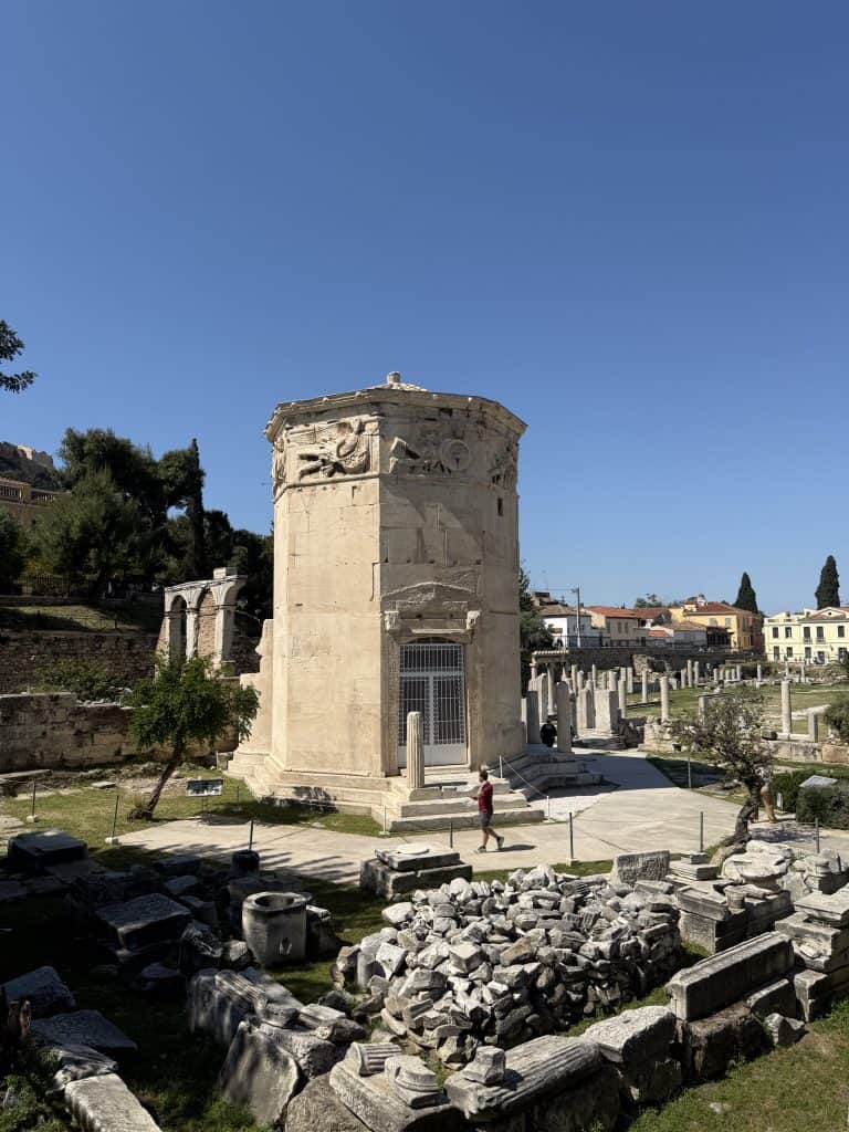 Close view of the Tower of the Winds in the Roman Agora of Athens, an octagonal marble structure decorated with carved reliefs near the roof. Broken stone blocks and archaeological remains surround the tower under a deep blue sky.