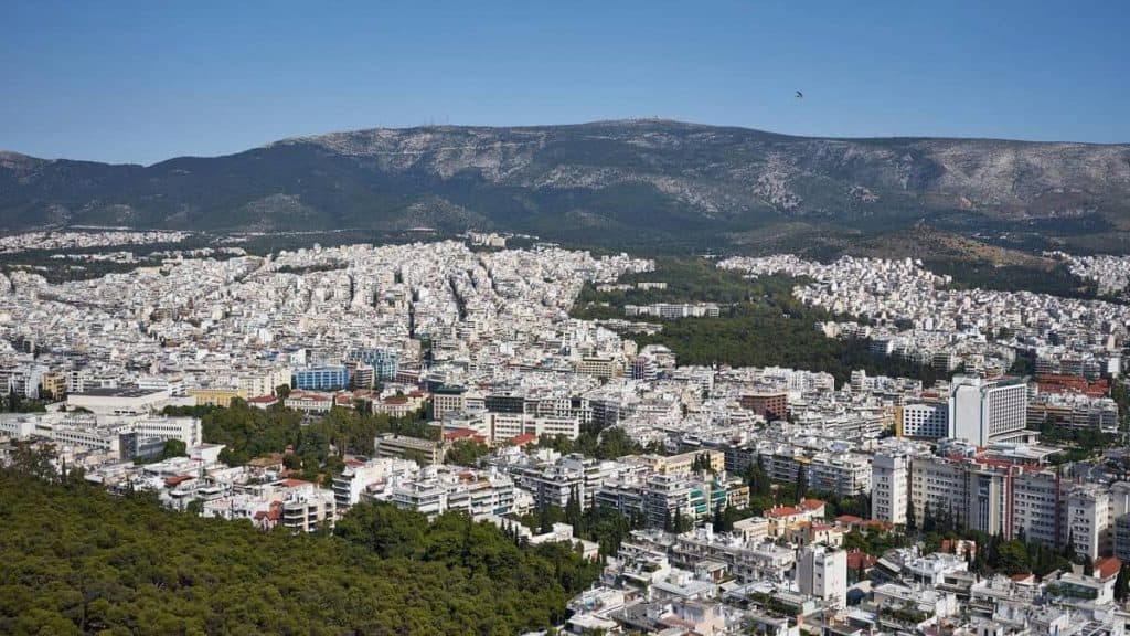 Panoramic view of Athens spreading across the foothills of Mount Hymettus, with dense white apartment buildings, green patches of trees, and a clear blue summer sky seen from Lycabettus Hill.