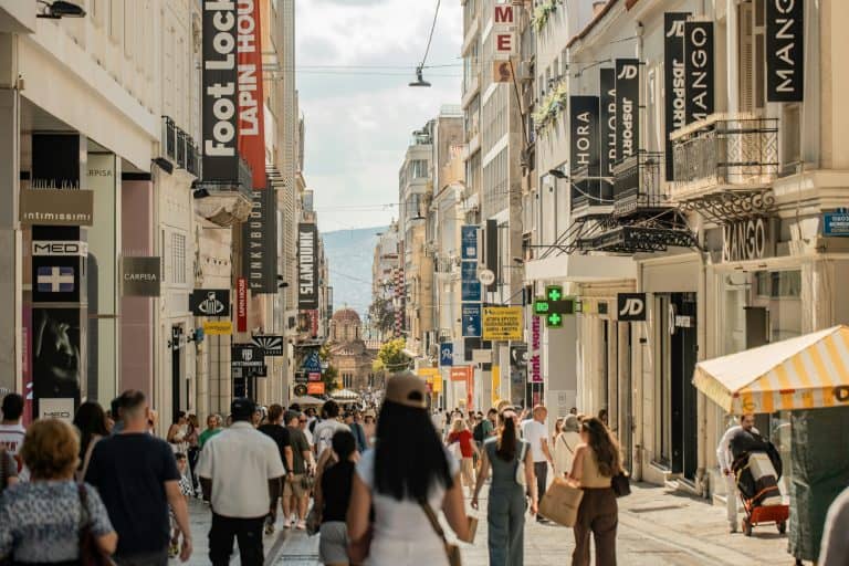 The busy pedestrian Ermou shopping street lined with storefronts and signs, with crowds walking toward a domed church and hills in the distance.