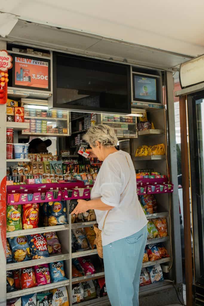A woman browses snacks at a small shop kiosk, surrounded by shelves and colorful displays of chips, candy, and other packaged foods.